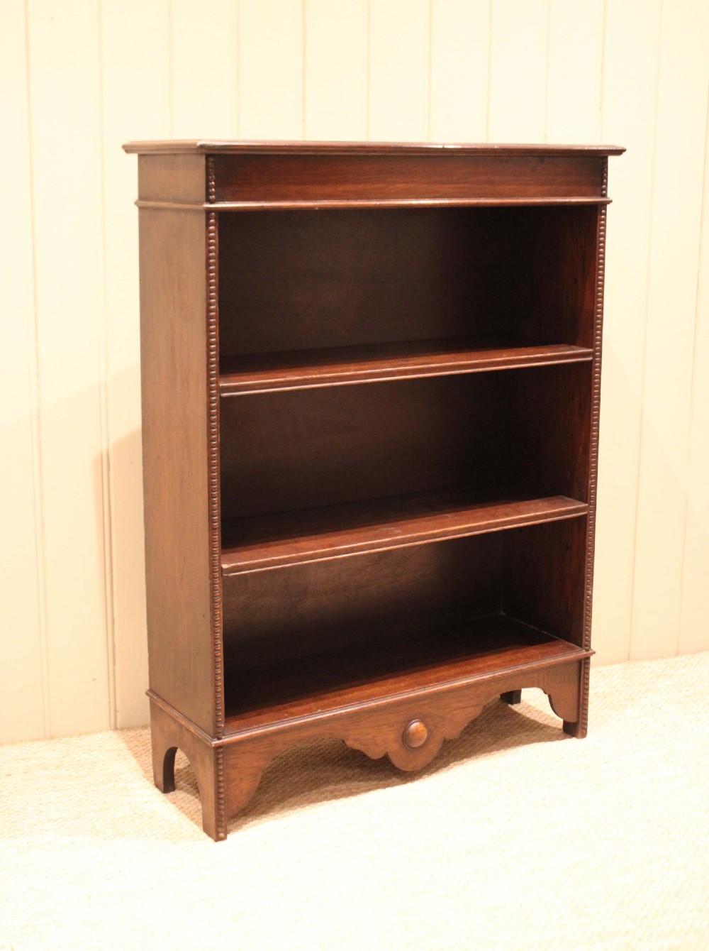 A dark oak open bookcase featuring three adjustable shelves, beaded moulding along the side columns, and a decorative shaped bottom apron with a central circular boss. View VIII