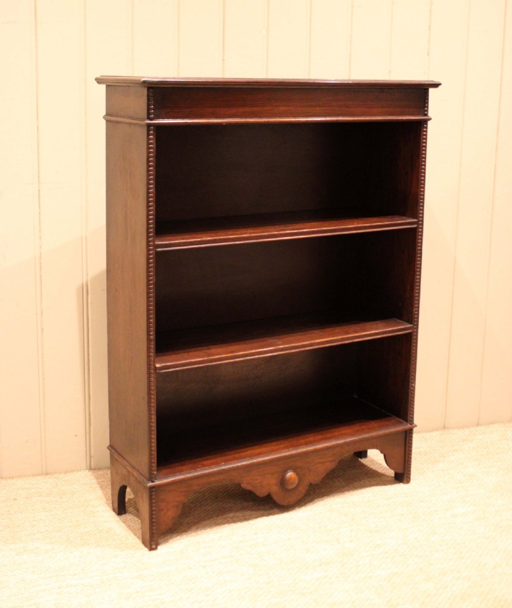 A dark oak open bookcase featuring three adjustable shelves, beaded moulding along the side columns, and a decorative shaped bottom apron with a central circular boss. View X