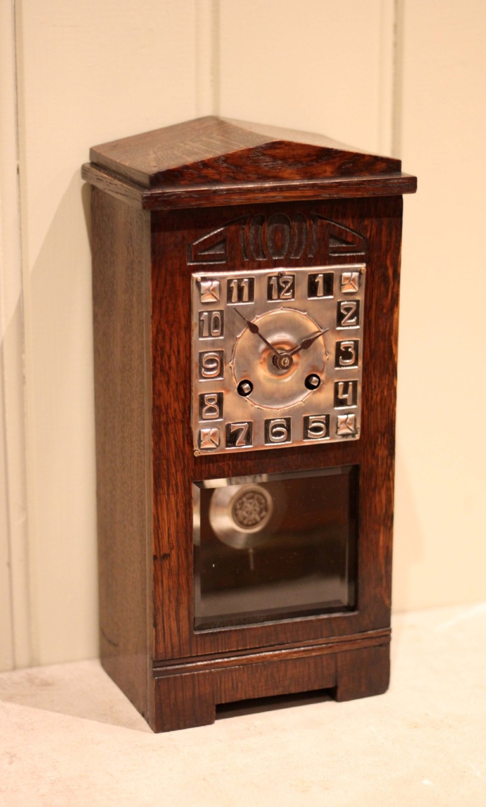 An Arts and Crafts dark oak mantel clock with a pediment top, carved geometric detailing, a square silvered metal dial, and a lower glass aperture displaying the pendulum.