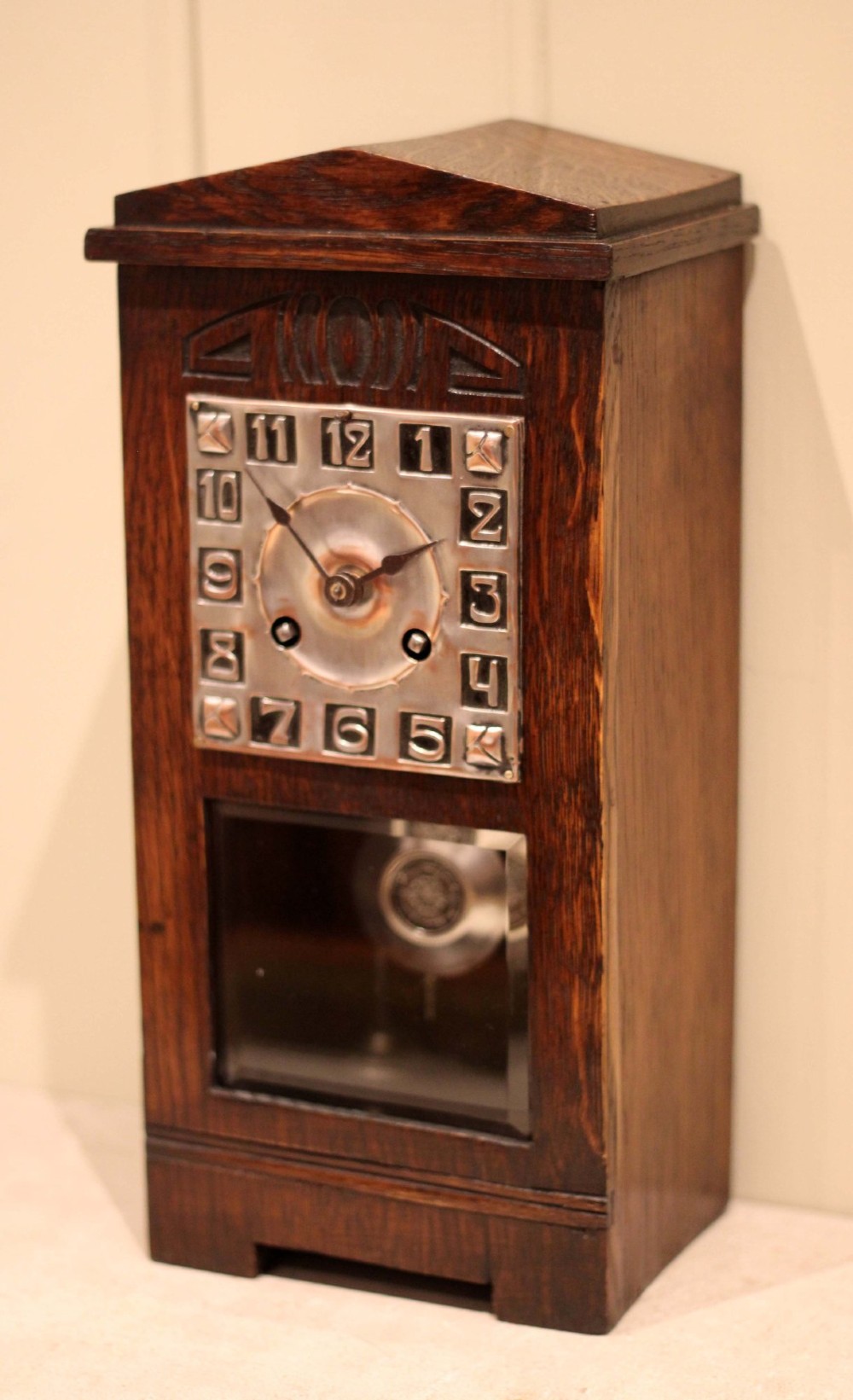 An Arts and Crafts dark oak mantel clock with a pediment top, carved geometric detailing, a square silvered metal dial, and a lower glass aperture displaying the pendulum. View III