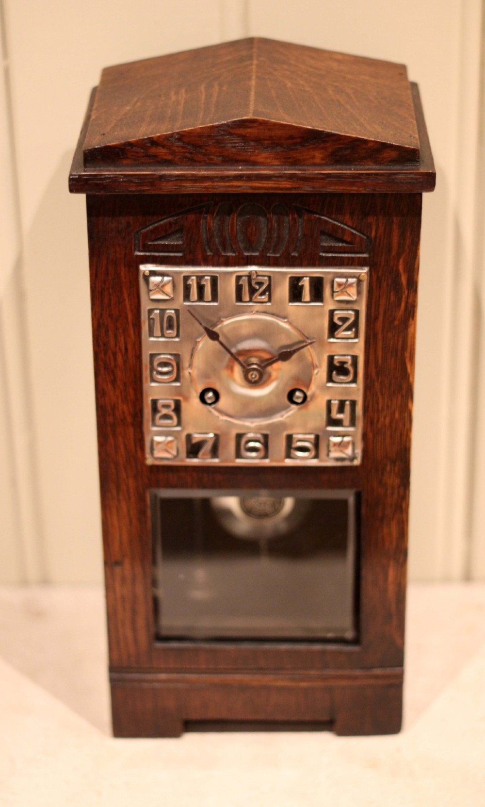An Arts and Crafts dark oak mantel clock with a pediment top, carved geometric detailing, a square silvered metal dial, and a lower glass aperture displaying the pendulum. View V