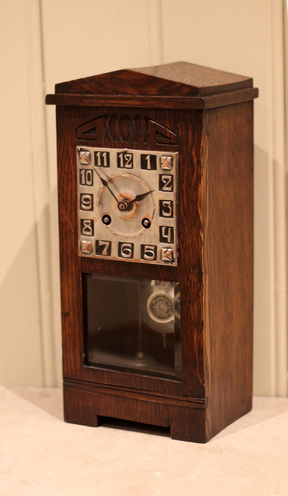 An Arts and Crafts dark oak mantel clock with a pediment top, carved geometric detailing, a square silvered metal dial, and a lower glass aperture displaying the pendulum. View VI