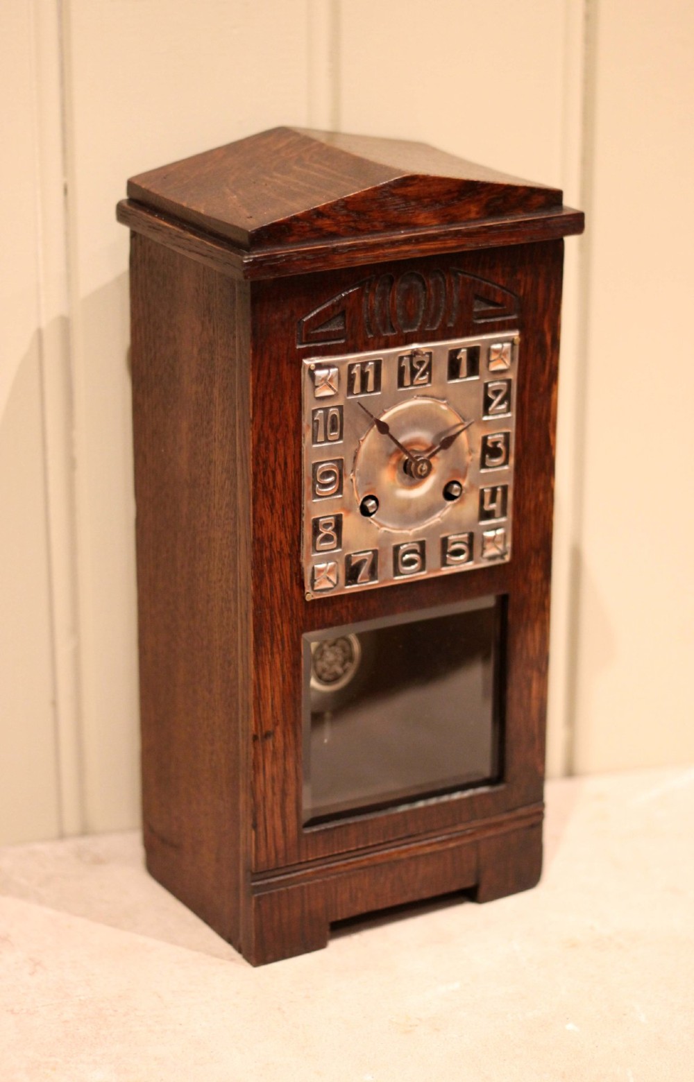 An Arts and Crafts dark oak mantel clock with a pediment top, carved geometric detailing, a square silvered metal dial, and a lower glass aperture displaying the pendulum. View VII