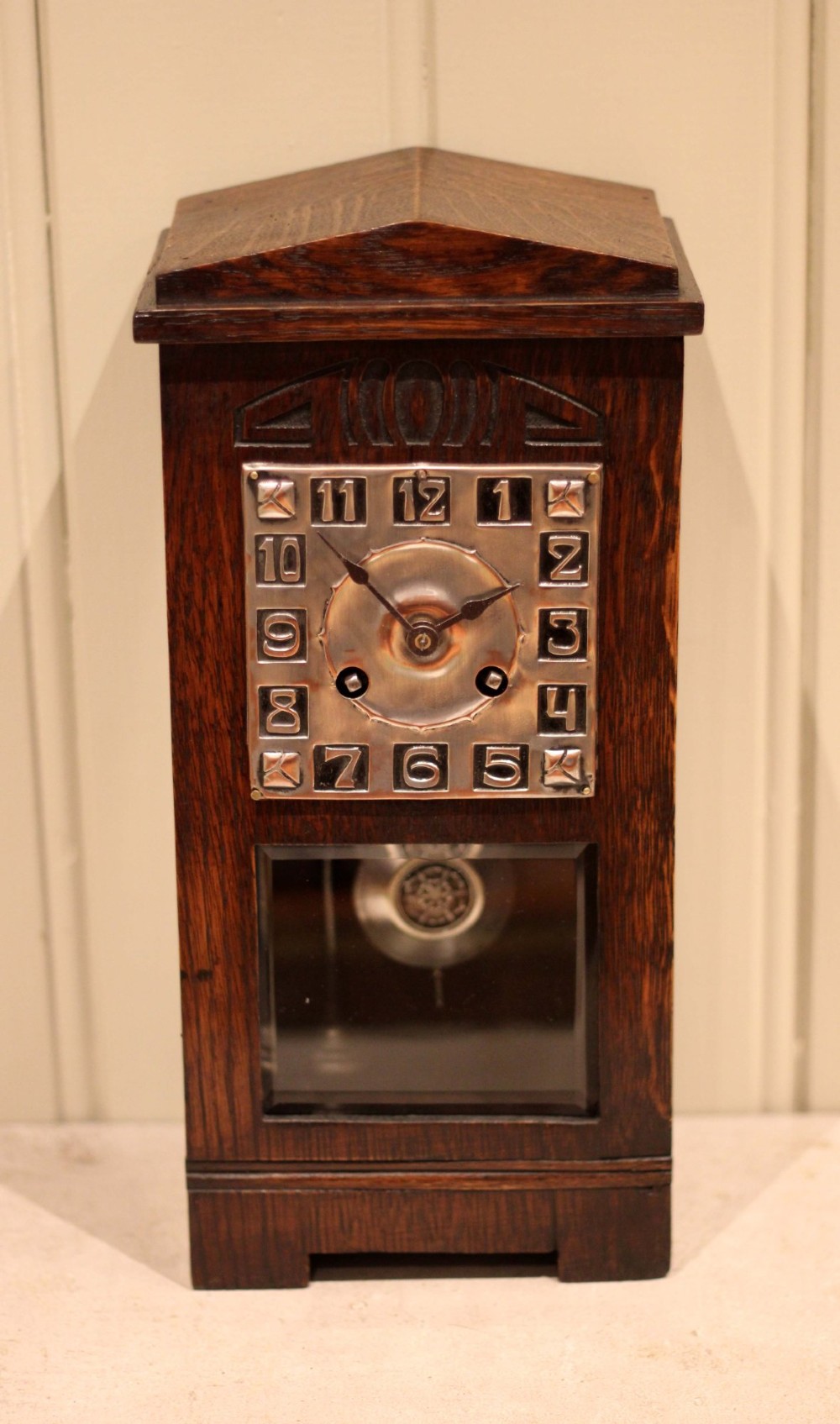 An Arts and Crafts dark oak mantel clock with a pediment top, carved geometric detailing, a square silvered metal dial, and a lower glass aperture displaying the pendulum. View VIII