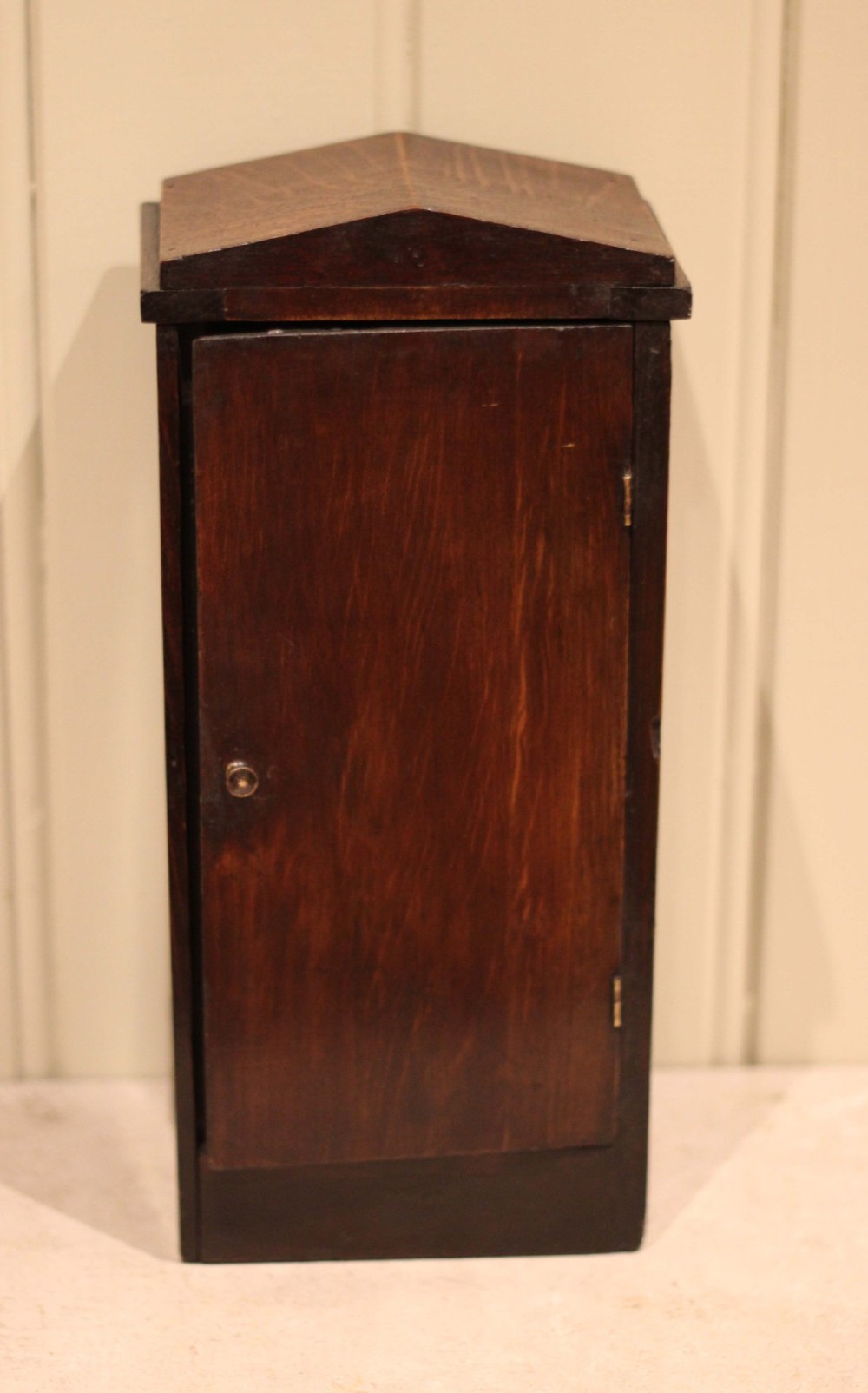 An Arts and Crafts dark oak mantel clock with a pediment top, carved geometric detailing, a square silvered metal dial, and a lower glass aperture displaying the pendulum. View IX