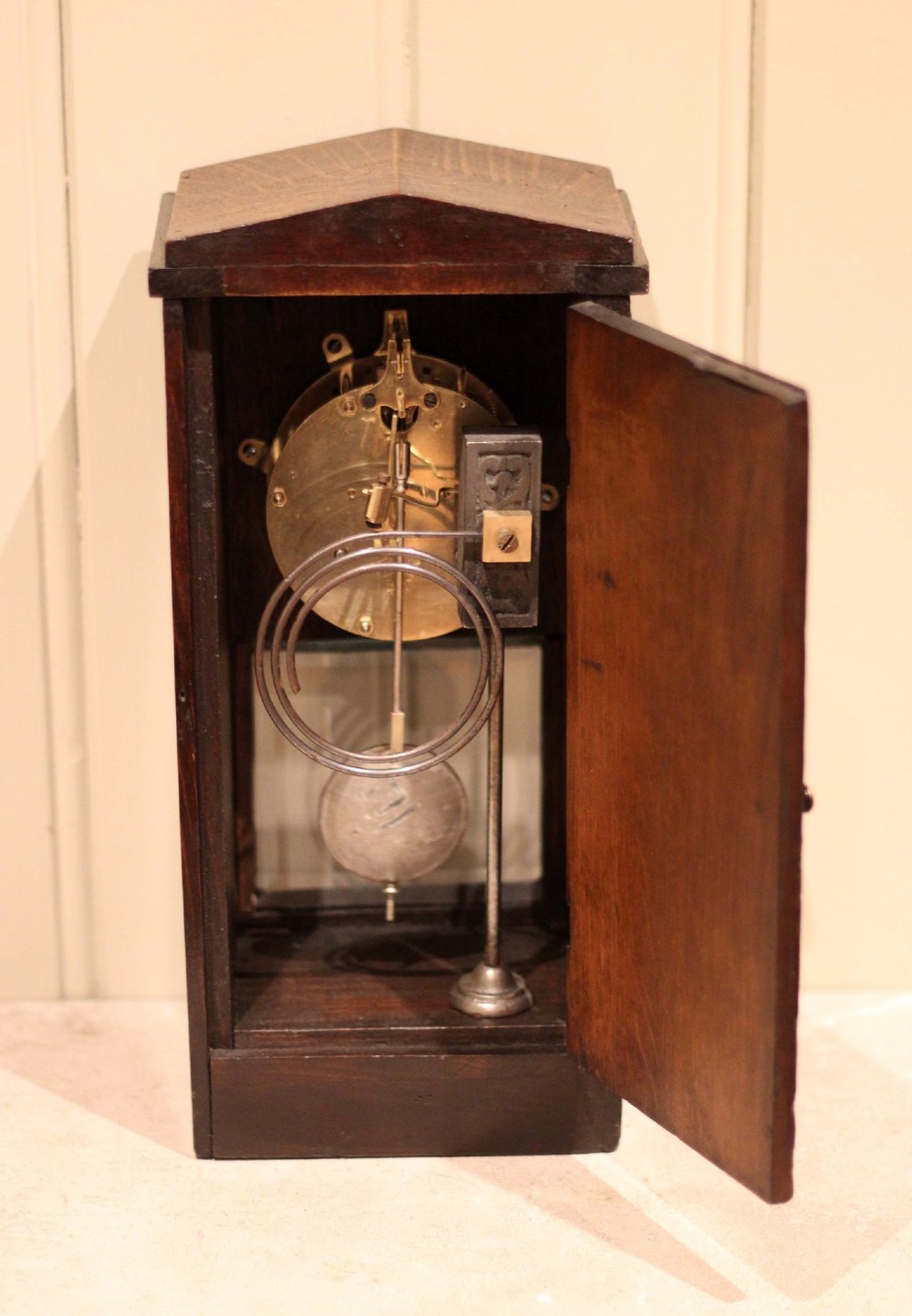 An Arts and Crafts dark oak mantel clock with a pediment top, carved geometric detailing, a square silvered metal dial, and a lower glass aperture displaying the pendulum. View X