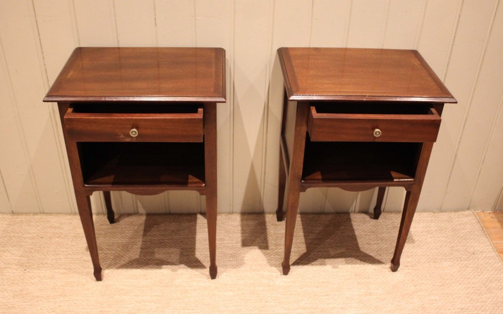 A matching pair of warm brown mahogany bedside tables, each featuring a single top drawer with a central brass knob pull above an open compartment, supported by slender tapered legs. View VI