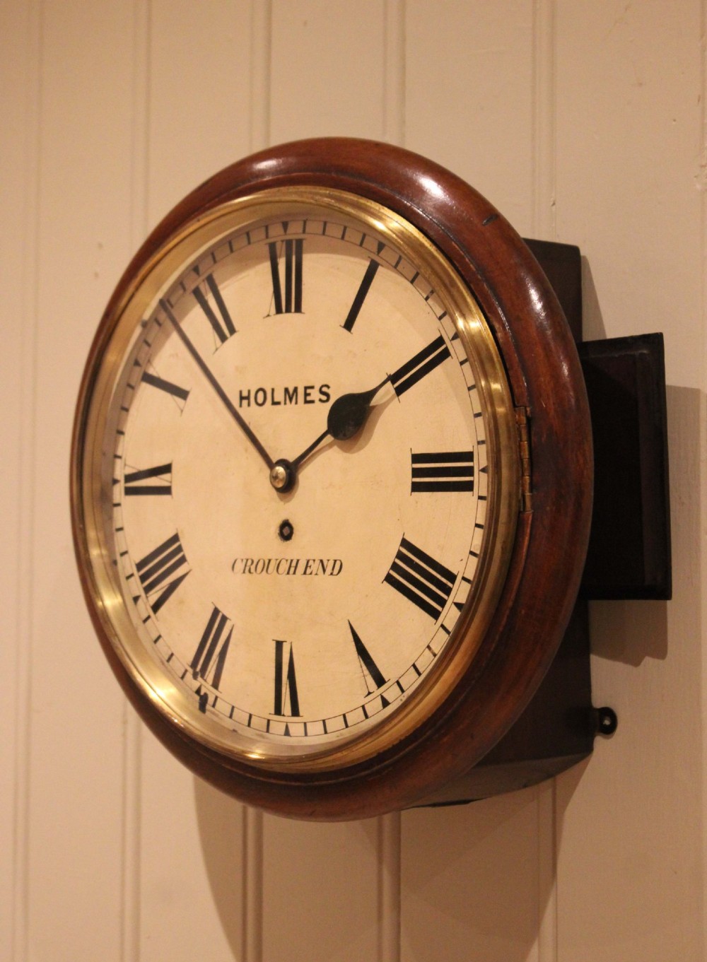A circular mahogany wall clock featuring a brass bezel surrounding an off-white dial with black Roman numerals. View III