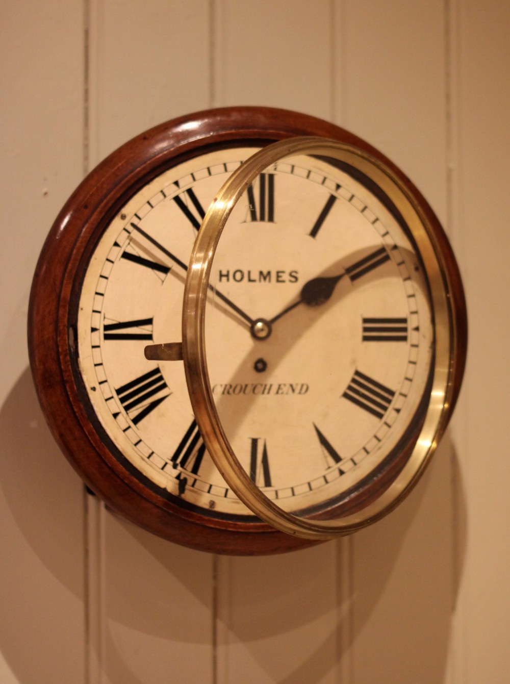 A circular mahogany wall clock featuring a brass bezel surrounding an off-white dial with black Roman numerals. View V