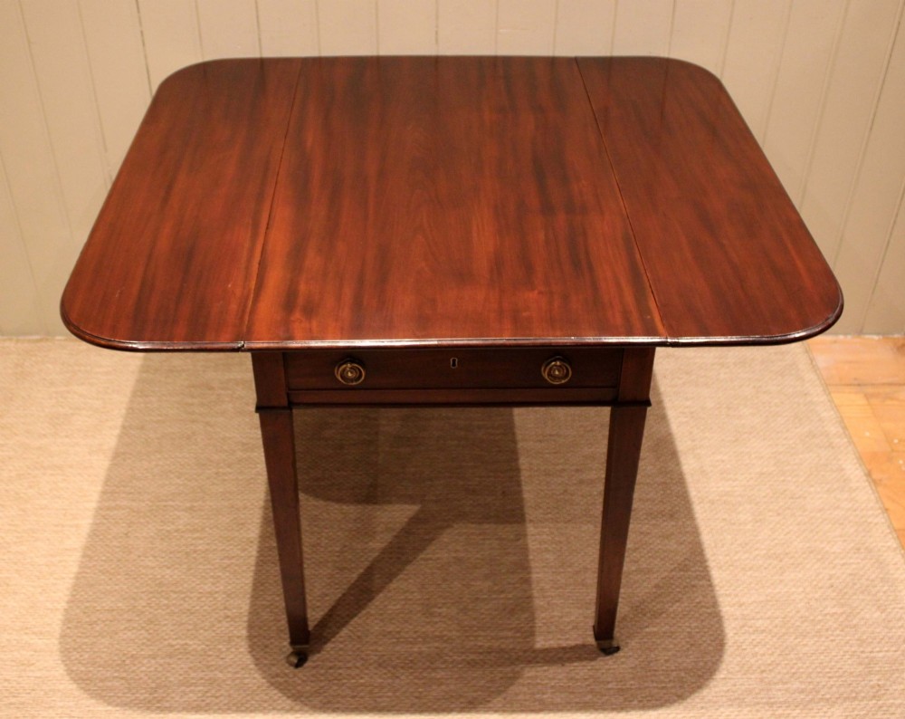 A solid mahogany Pembroke table featuring two drop leaves, a central drawer with brass ring handles, and square tapered legs fitted with brass castors. View II