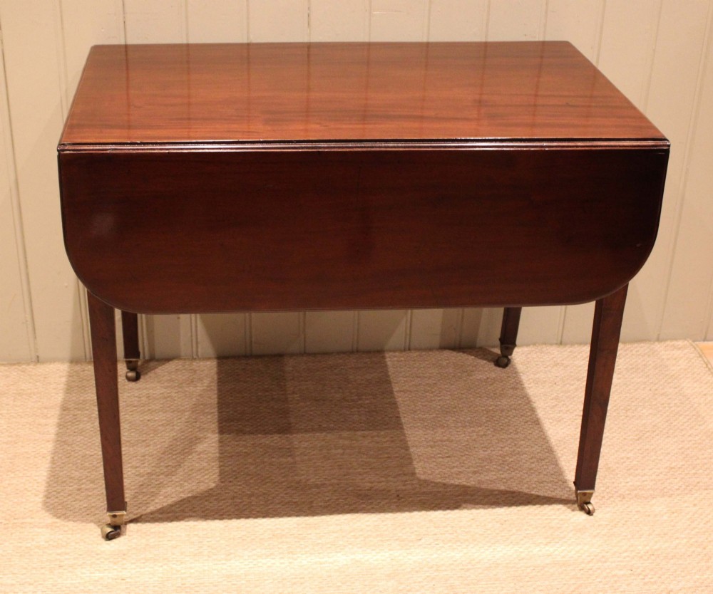 A solid mahogany Pembroke table featuring two drop leaves, a central drawer with brass ring handles, and square tapered legs fitted with brass castors. View III