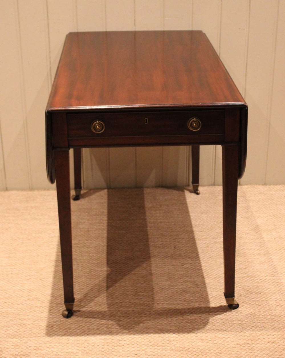 A solid mahogany Pembroke table featuring two drop leaves, a central drawer with brass ring handles, and square tapered legs fitted with brass castors. View IV