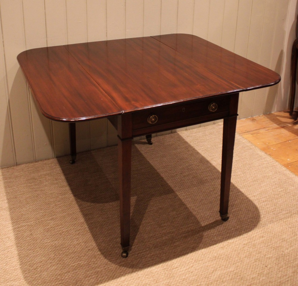 A solid mahogany Pembroke table featuring two drop leaves, a central drawer with brass ring handles, and square tapered legs fitted with brass castors. View V