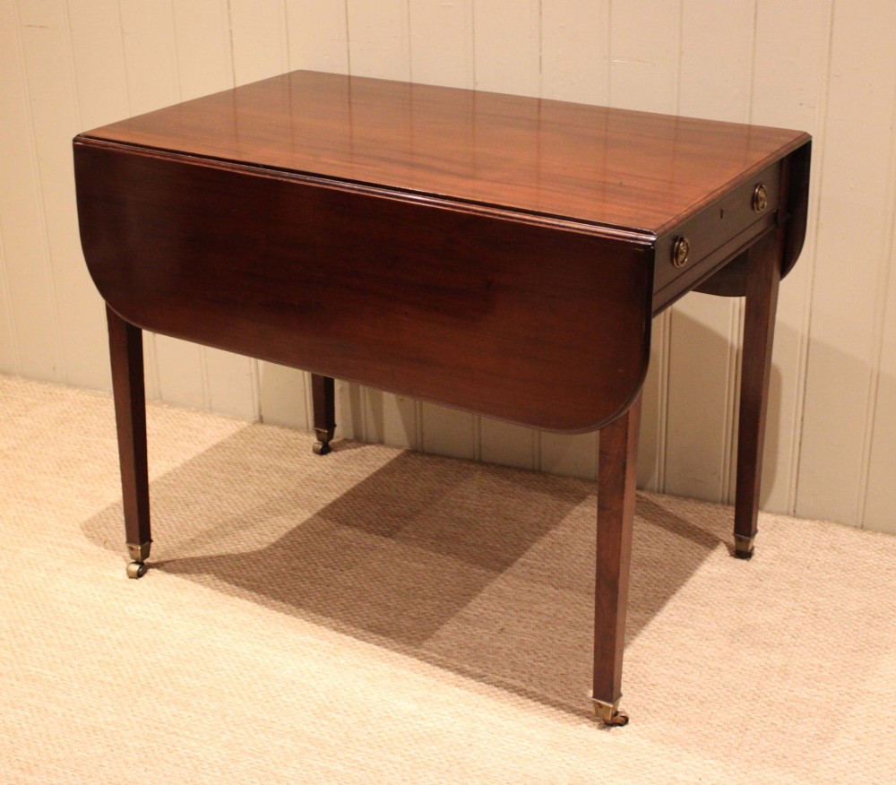A solid mahogany Pembroke table featuring two drop leaves, a central drawer with brass ring handles, and square tapered legs fitted with brass castors. View VI