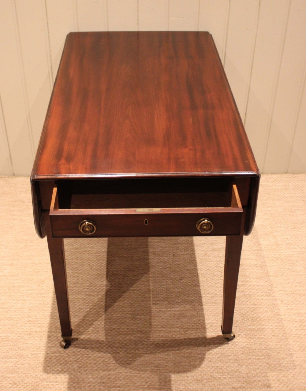 A solid mahogany Pembroke table featuring two drop leaves, a central drawer with brass ring handles, and square tapered legs fitted with brass castors. View VII