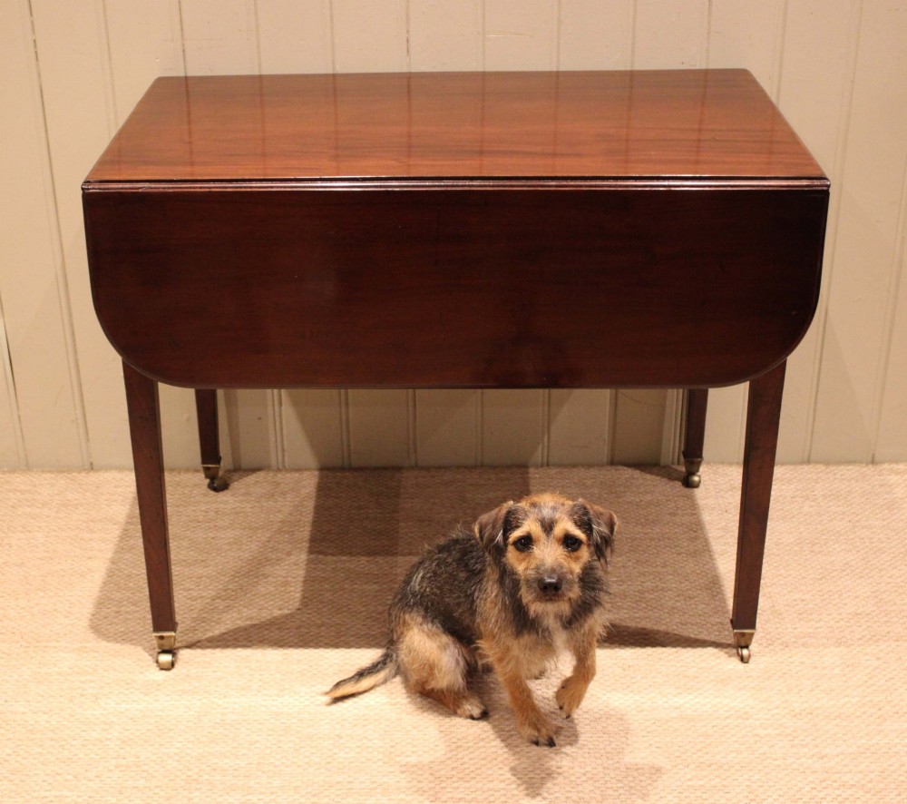 A solid mahogany Pembroke table featuring two drop leaves, a central drawer with brass ring handles, and square tapered legs fitted with brass castors. View IX