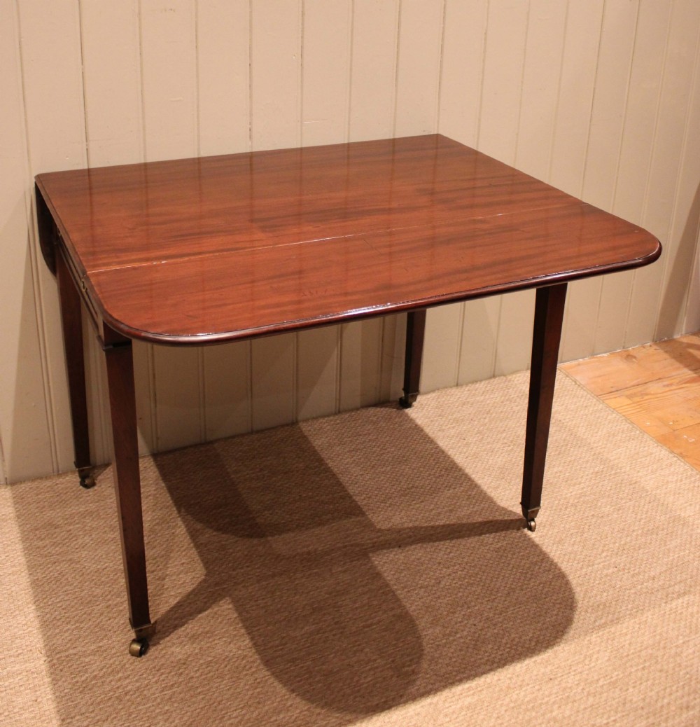 A solid mahogany Pembroke table featuring two drop leaves, a central drawer with brass ring handles, and square tapered legs fitted with brass castors. View X