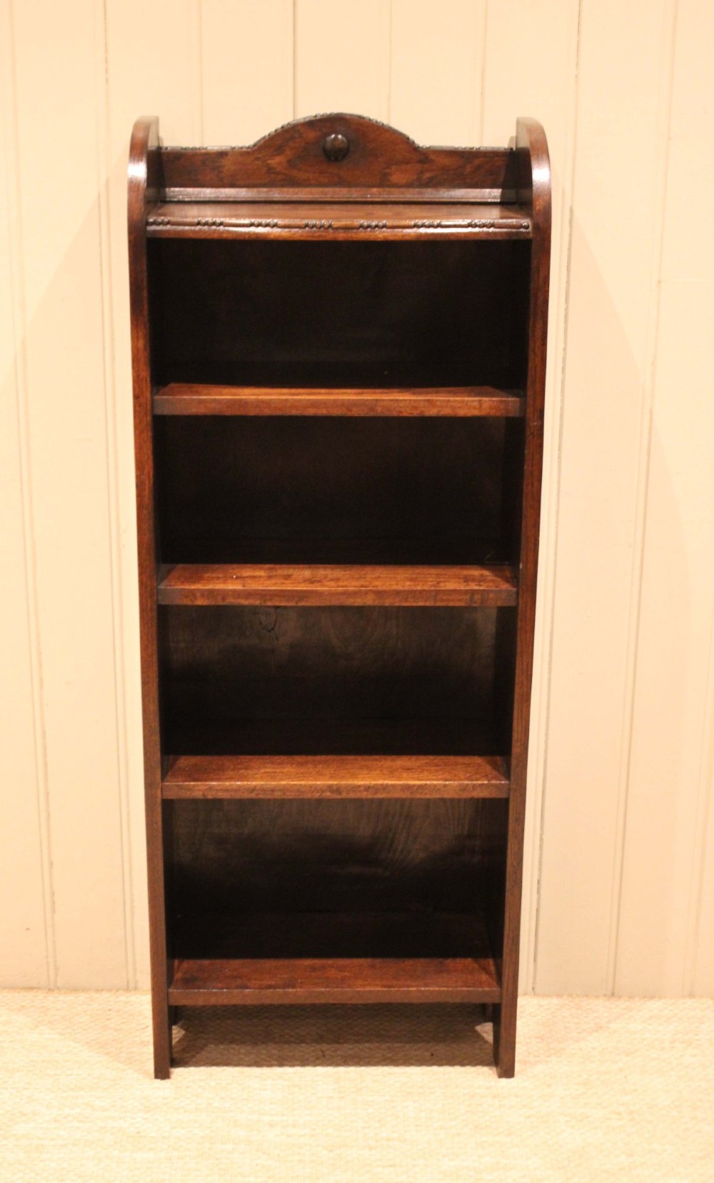 A slim freestanding dark oak open bookcase featuring four shelves and a shaped decorative pediment with a round motif. View II