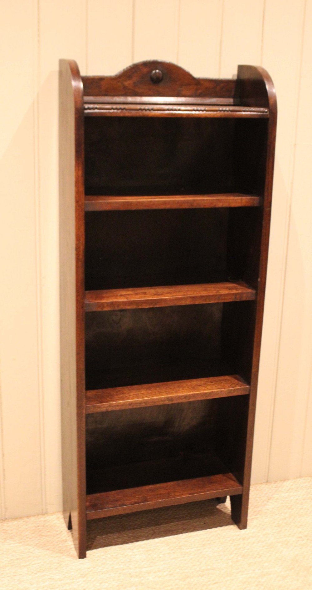 A slim freestanding dark oak open bookcase featuring four shelves and a shaped decorative pediment with a round motif. View IV