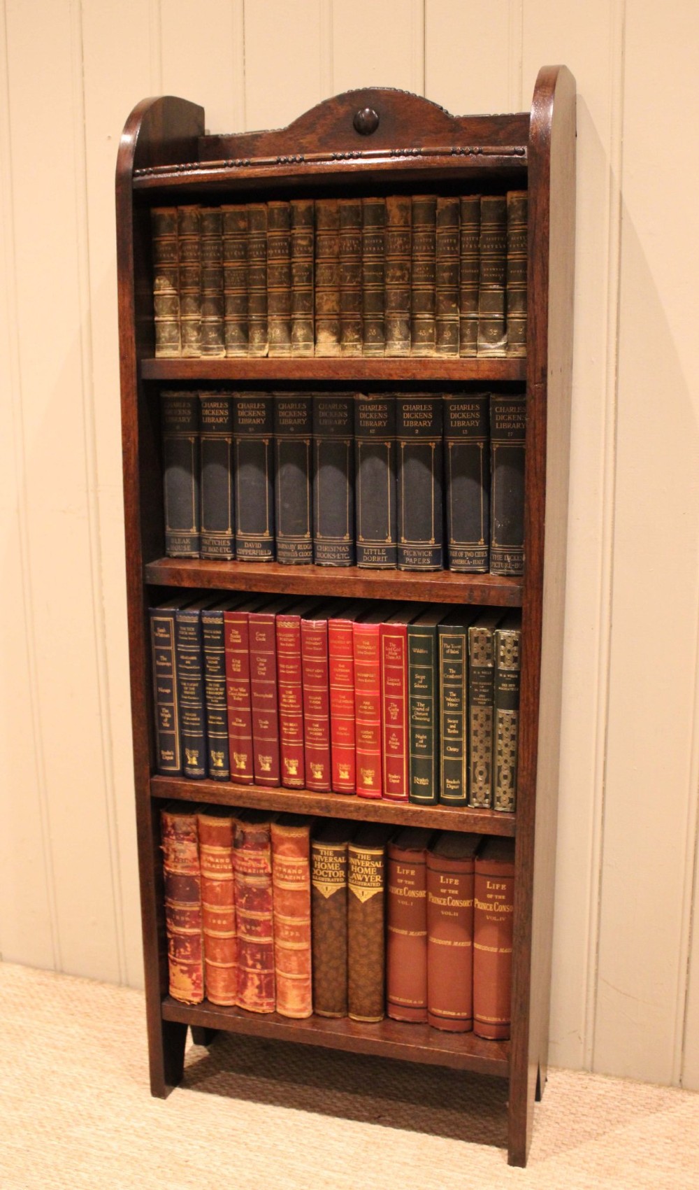 A slim freestanding dark oak open bookcase featuring four shelves and a shaped decorative pediment with a round motif. View V