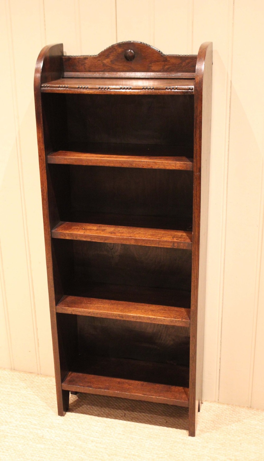 A slim freestanding dark oak open bookcase featuring four shelves and a shaped decorative pediment with a round motif. View VI