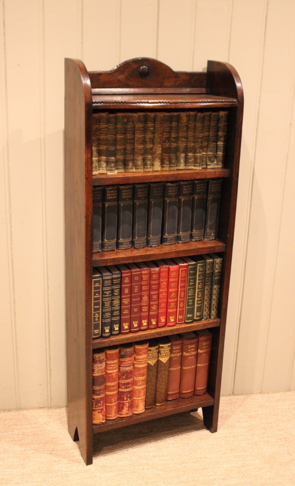 A slim freestanding dark oak open bookcase featuring four shelves and a shaped decorative pediment with a round motif. View VII