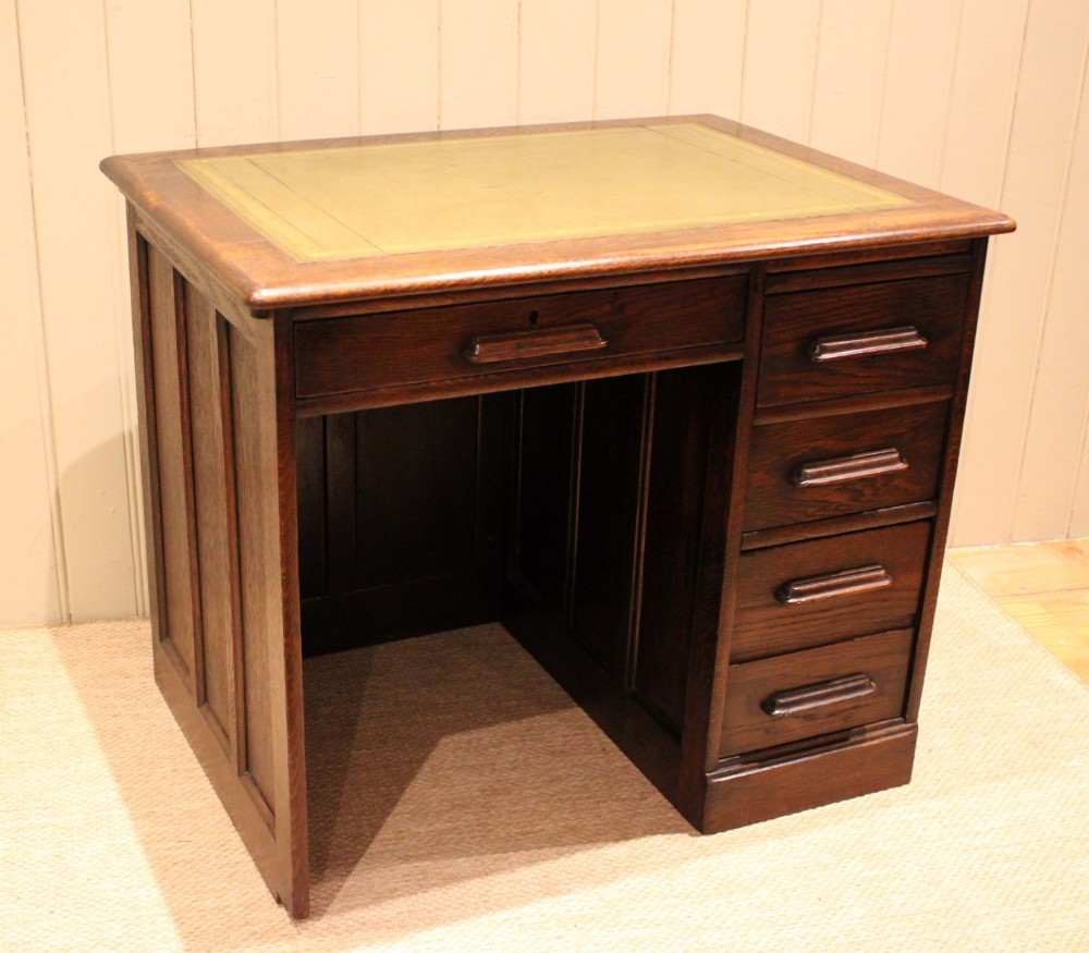 A small solid dark oak clerks desk featuring a leather inset writing surface, a single central drawer, and a four-drawer pedestal on the right.