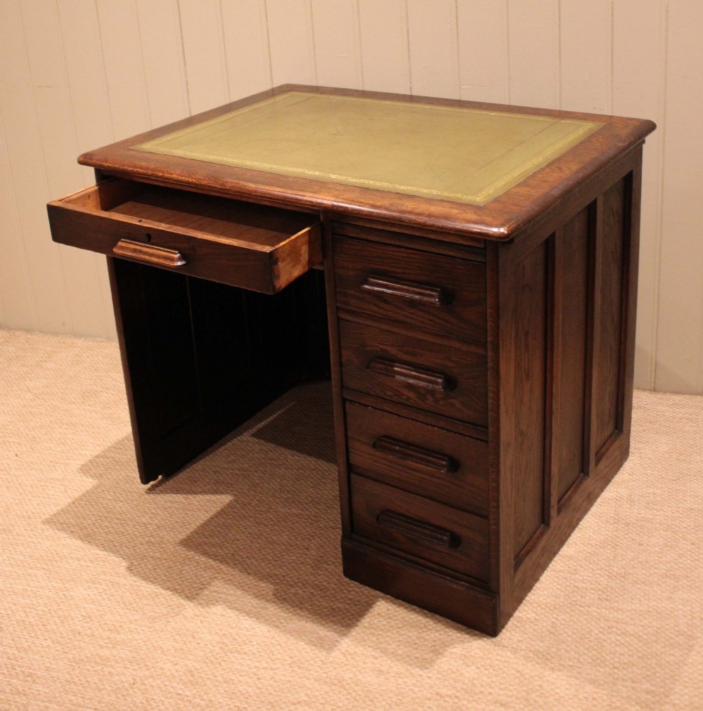 A small solid dark oak clerks desk featuring a leather inset writing surface, a single central drawer, and a four-drawer pedestal on the right. View V