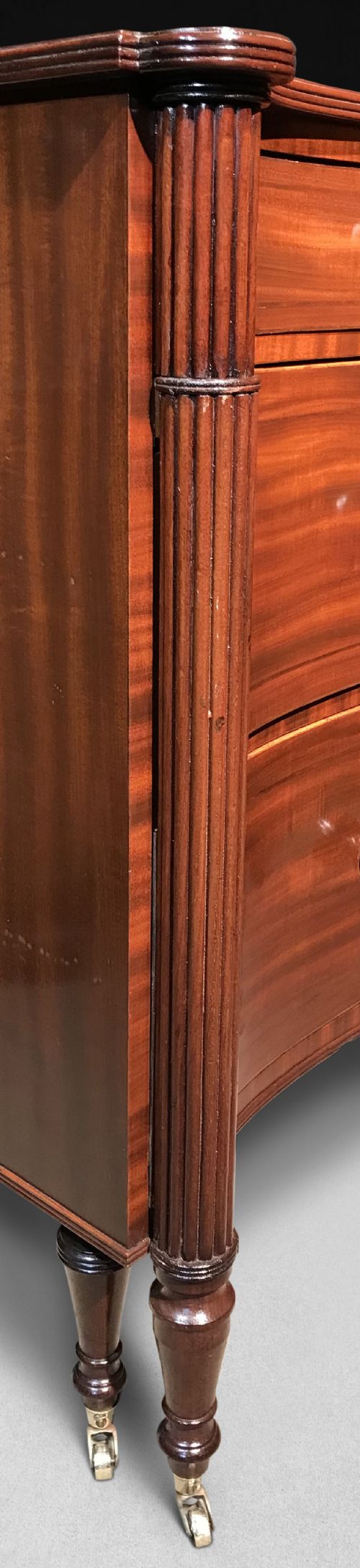 fig 3. A Regency mahogany concave-fronted commode featuring three drawers, ebony line inlay, reeded side columns, turned legs, and brass castors.