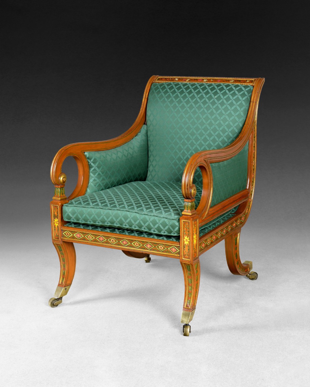 A Regency period library armchair constructed from painted and gilt-simulated satinwood with scrolled arms, upholstered in patterned green fabric and supported on brass castors.