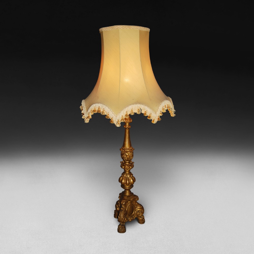 A 19th-century Italian carved giltwood table lamp featuring an ornate baluster stem, scrolling tripod base, and a cream fabric lampshade with fringed trim.