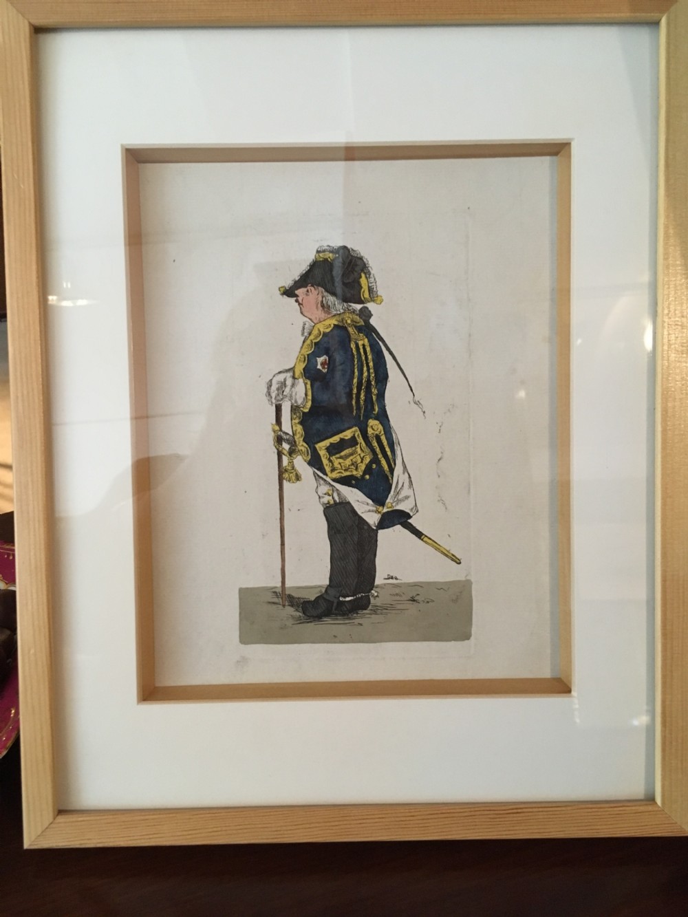 A framed hand-coloured Georgian engraving depicting a military officer in profile, wearing a blue coat with gold trim, a tricorn hat, and boots, mounted within a light wood frame.