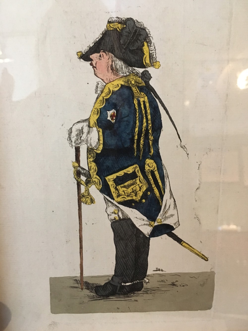 A framed hand-coloured Georgian engraving depicting a military officer in profile, wearing a blue coat with gold trim, a tricorn hat, and boots, mounted within a light wood frame. View II