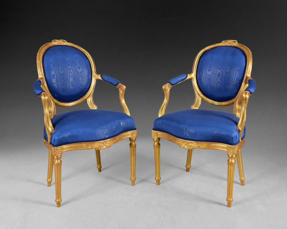 A pair of George III carved giltwood salon armchairs featuring oval backrests, padded arms, fluted legs, and blue silk upholstery.