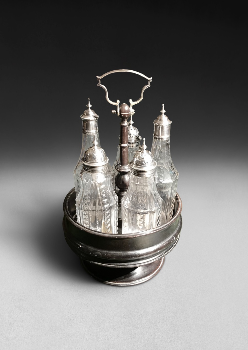 An ebonised wooden circular cruet stand holding five cut glass bottles with fitted silver caps and a central carrying handle.