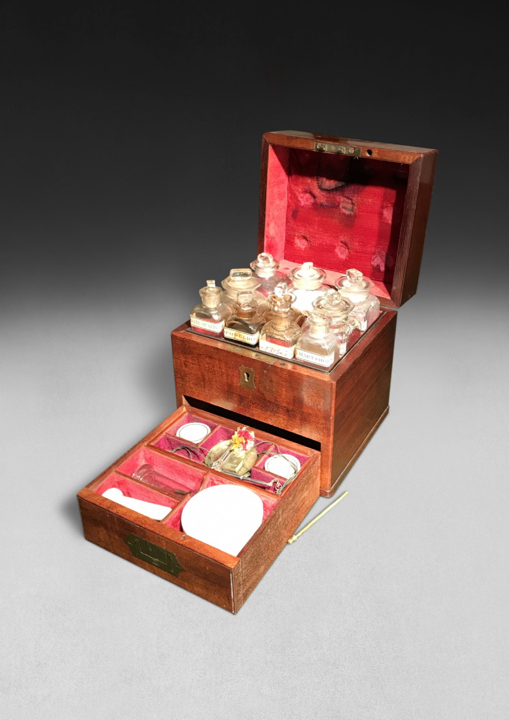 An open Regency mahogany apothecary box featuring glass medicine phials in the top section and a fitted lower drawer lined in red fabric containing period tools.