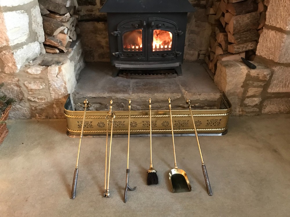 A polished brass fireplace fender featuring pierced geometric cut-outs and rope-twist trim, displayed alongside a six-piece brass fire tool set.