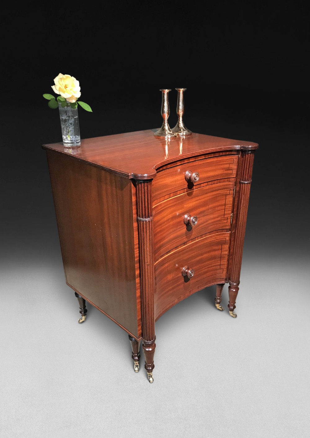 A Regency mahogany concave-fronted commode featuring three drawers, ebony line inlay, reeded side columns, turned legs, and brass castors.