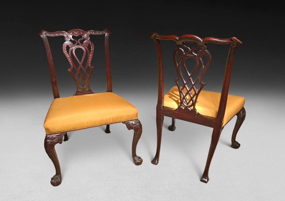 A pair of Chippendale style mahogany dining chairs featuring intricate carved ribbon back splats, yellow upholstered seats with brass studs, and front cabriole legs with claw and ball feet.