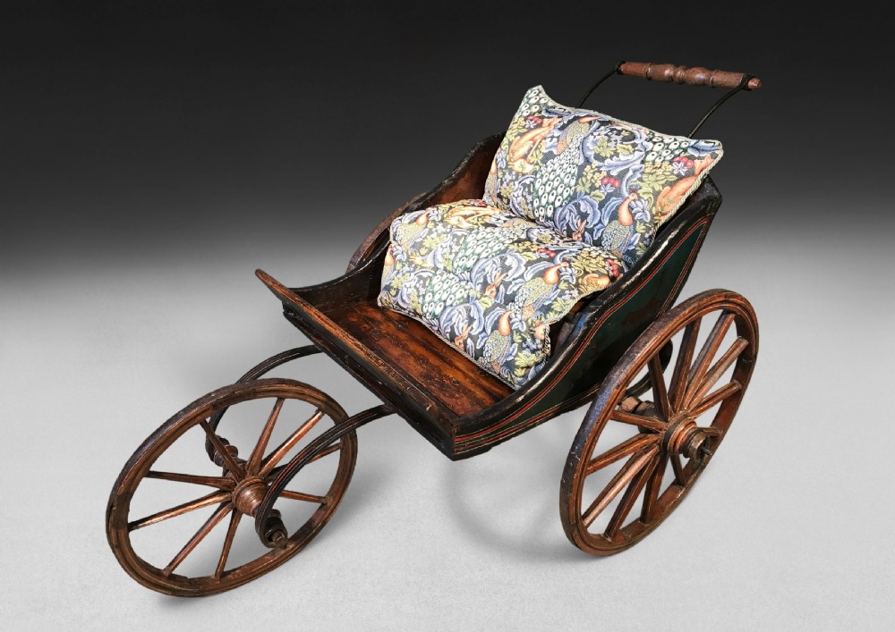 An early 19th-century three-wheeled wooden childs pushchair featuring hand-painted polychrome details, large spoked wheels, a turned handle, and patterned cushions.