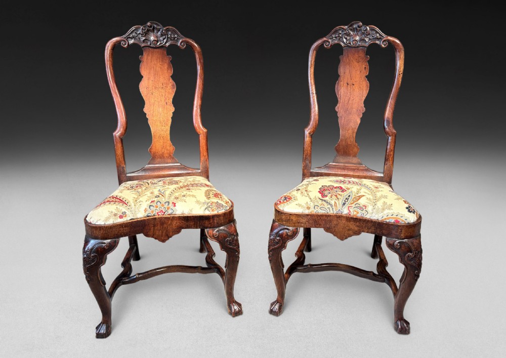 A pair of antique George I walnut side chairs featuring carved cresting rails, vase-shaped back splats, floral upholstered drop-in seats, and cabriole front legs.