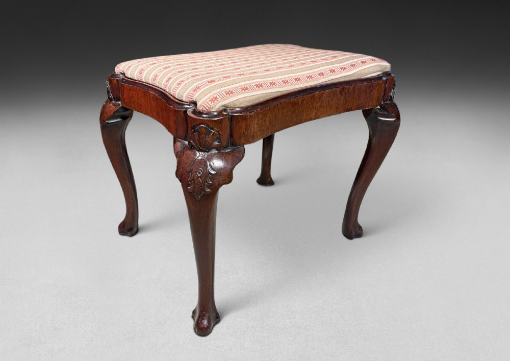 A mid-18th century mahogany stool featuring a serpentine upholstered seat, carved cabriole legs, and shell motifs on the knees.