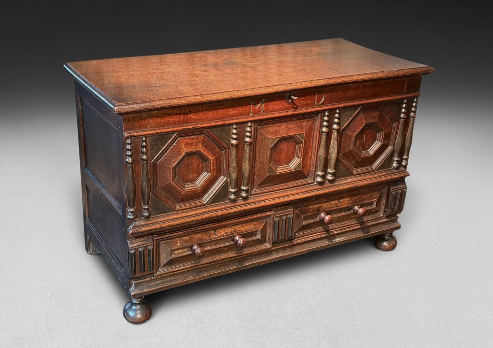An 18th-century dark oak mule chest featuring geometric carved panels, split spindle turnings, two base drawers, and turned bun feet.