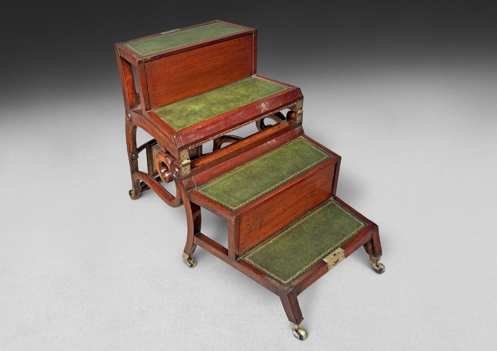 A Regency mahogany metamorphic armchair featuring scrolled arms, a cane back panel, a green leather seat cushion, and brass castors that transforms into library steps. View V