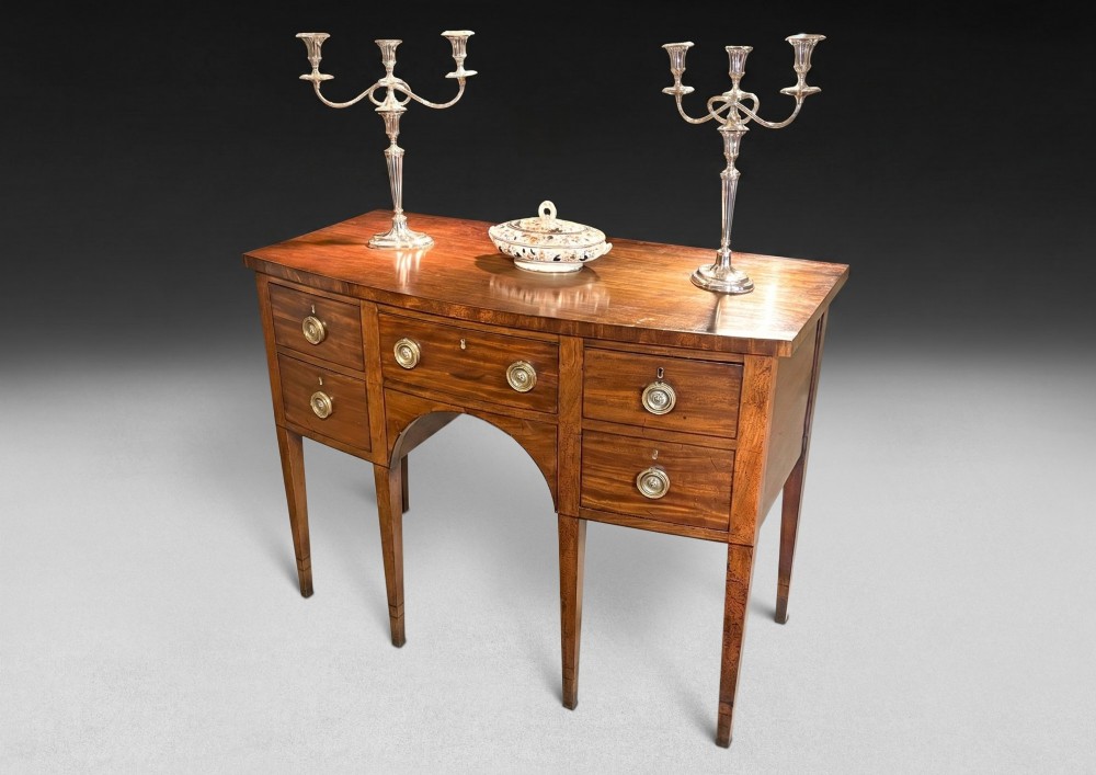 A George III mahogany bowfront sideboard featuring five drawers with circular brass ring handles, an arched central apron, and four square tapered legs. View II