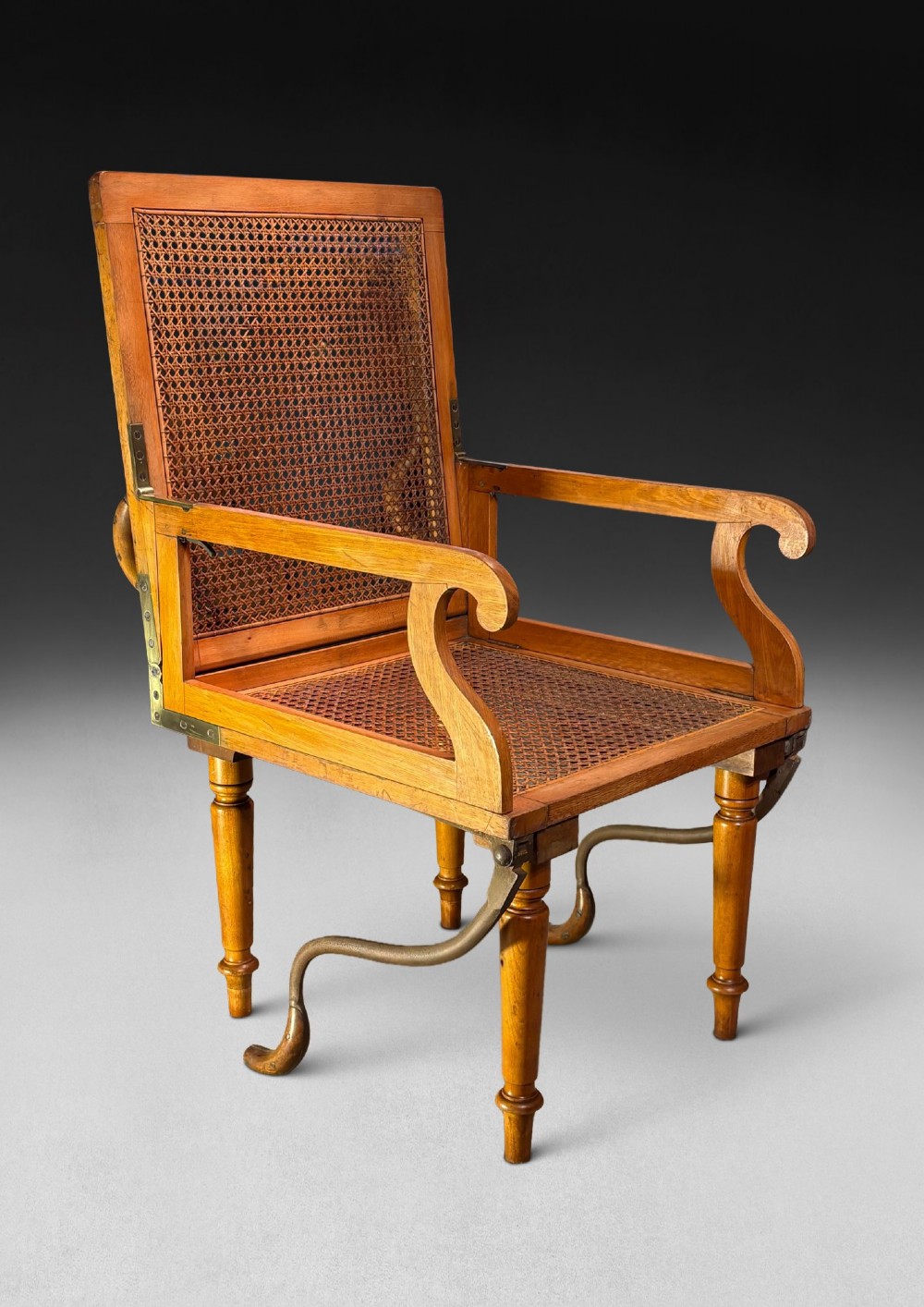 A mid-19th-century wooden campaign armchair with a woven cane back and seat, brass corner mounts, turned legs, and extendable curved handles. View II