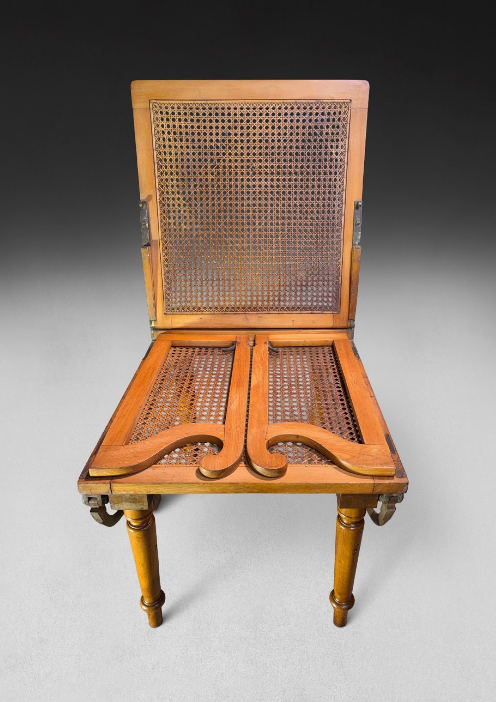 A mid-19th-century wooden campaign armchair with a woven cane back and seat, brass corner mounts, turned legs, and extendable curved handles. View V