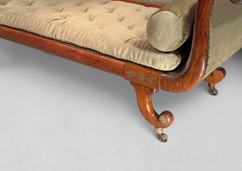 Regency simulated rosewood chaise longue with brass inlay, featuring asymmetrical scroll ends, turned brass-castor legs, olive green button-tufted upholstery, and matching bolster cushions. View II