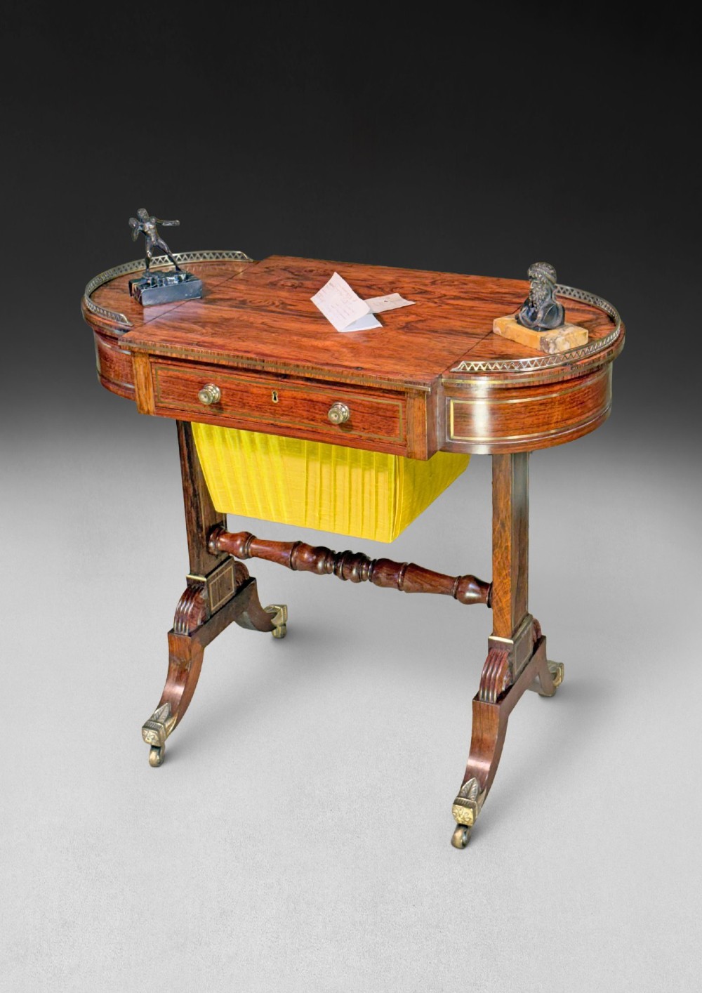 A Regency rosewood compendium table with brass stringing inlay, a single drawer, a pleated yellow silk basket underneath, and splayed legs ending in brass castors.