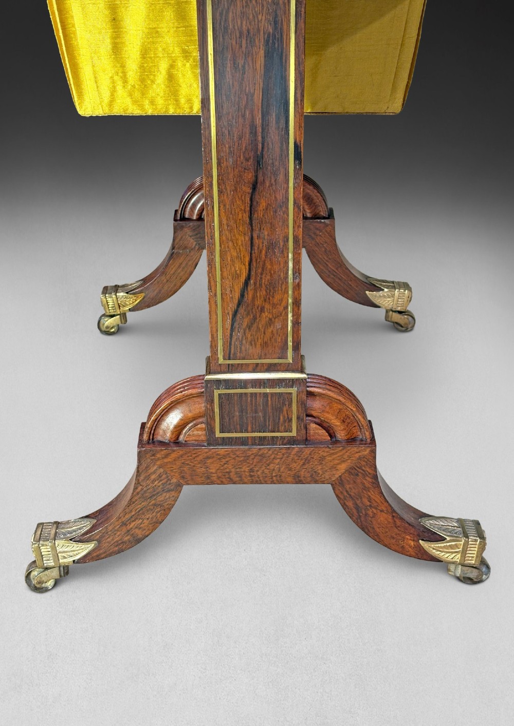 A Regency rosewood compendium table with brass stringing inlay, a single drawer, a pleated yellow silk basket underneath, and splayed legs ending in brass castors. View II
